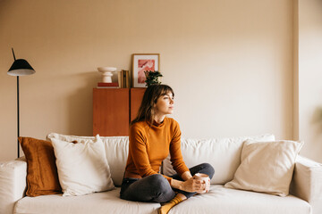 Obraz premium Woman sitting cross-legged on a white sofa contemplating while enjoying a hot drink