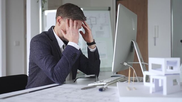 Businessman stressing at his office desk, covering face with hands. Feeling tired, having a headache from work overload. Business and architecture concept