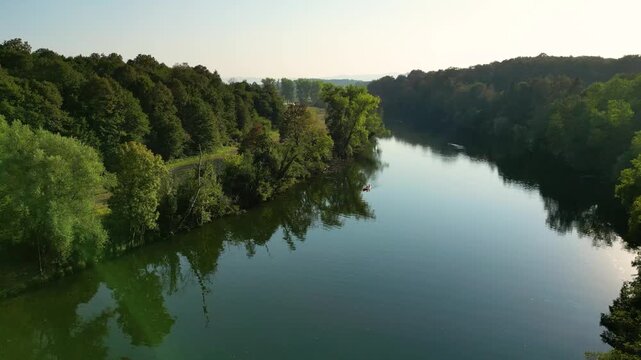 Aerial view of the Kolpa River near Metlika, Slovenia, with a lone canoeist paddling through calm water surrounded by lush green forest, reflected in the river during warm summer light.