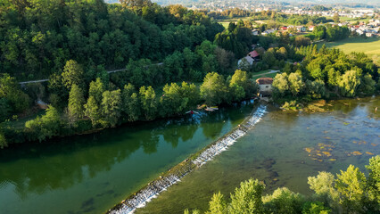 Fototapeta premium Aerial view of the Kolpa River near Metlika, Slovenia, featuring a small river dam with flowing water, calm green river pools, surrounding forest, and a peaceful rural landscape in summer.