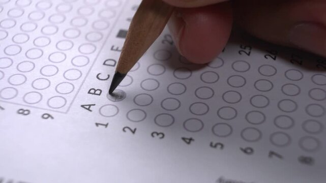 Close-up of a hand with a pencil shading in answer circles on a multiple choice test sheet or official form document