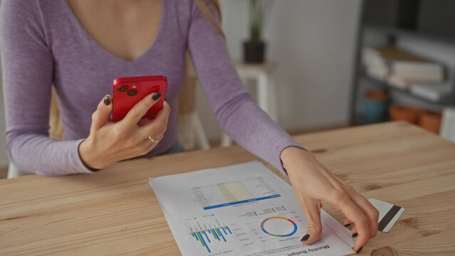 Woman holding smartphone and document in living room while analyzing data, surrounded by credit card and paperwork on wooden table, suggesting financial planning at home.