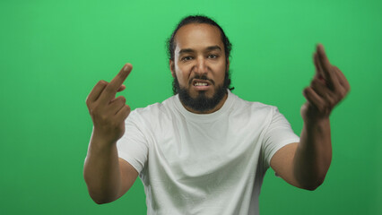 Naklejka premium Man with beard and white tshirt grimacing and shows middle fingers with both hands toward camera in a green studio; anger defiance.