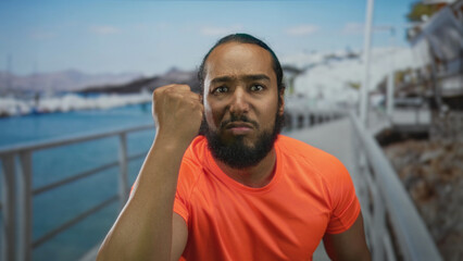 Fototapeta premium Man in orange shirt with hand to mouth gesture and clenched fist on a seaside street promenade near a pier; frustration confrontation challenge.