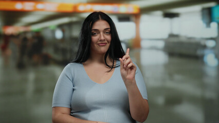 Fototapeta premium Woman gestures denying while pointing at herself in a busy airport terminal setting, expressing curiosity and conveying non-verbal communication in a bustling indoor environment.