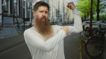 Fototapeta premium Bearded man in white shirt flexes bicep and grips muscle on city street with parked bicycles and trees; confidence.