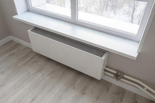white radiator mounted under a double-pane window with a white sill. Light wood laminate flooring, neutral walls, with visible plumbing pipes below the radiator. Winter view outside.