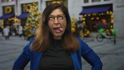 Fototapeta premium Woman wearing glasses and a blue jacket sticks out tongue on street with festive shop decorations; playfulness.