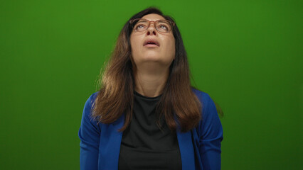 Fototapeta premium Hispanic woman raising hands toward ceiling and tilting head back in wide green studio set; frustration.