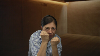 Fototapeta premium Hispanic woman with fists raised in studio against wooden panel wall, mid gesture with focused expression; determination.
