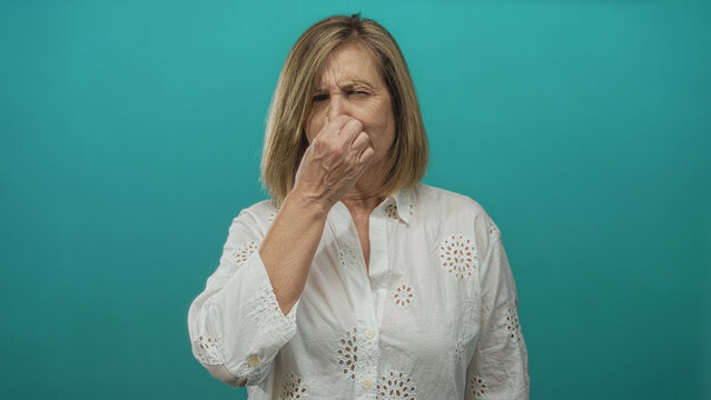 Middle aged caucasian woman grimaces and pinches her nose in teal studio setting; disgust odor stink.
