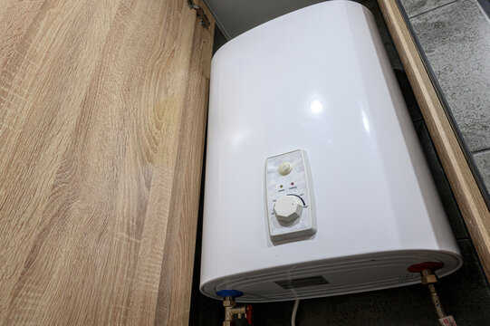 white electric storage water heater mounted under a wooden cabinet, featuring a control panel with a knob and indicator lights, connected via blue and red pipes.