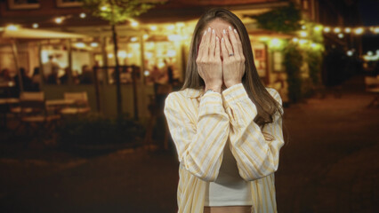 Woman covering eyes with hands on a restaurant terrace by a lit street in the evening, playful pose; playful surprise.