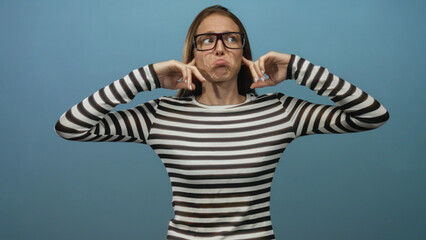 Fototapeta premium Woman in striped shirt and glasses plugs ears with fingers in blue studio setting while squinting and pursing lips; frustration defiance.