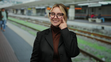 Woman at train station pointing to eye wearing glasses and black jacket with background of railway...