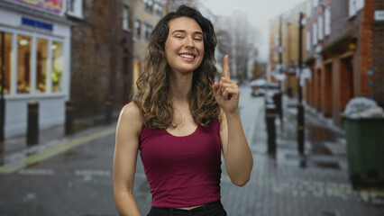 Woman in burgundy tanktop smiles and crosses bare arms on wet cobblestone street under soft daylight; confidence empowerment joy selfassurance.