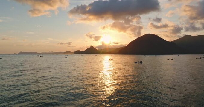 Drone flight over fishing boats floats on tranquil Selong Belanak Beach water, mountains in background, bright sunset sky with warm, golden light during a tropical evening in Lombok, Indonesia