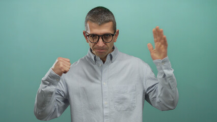 Naklejka premium Man standing against blue wall with fists up, wearing glasses and striped shirt, poised for fight, displaying confident expression and focus, suggesting readiness and anticipation.