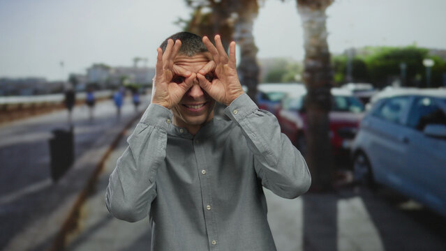 Man making binoculars gesture outdoors with a playful expression on a sunny street lined with palm trees and parked cars in the background.