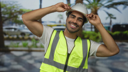 Naklejka premium Man adjusts white hardhat while wearing reflective vest on urban street lined with palm trees and parked cars; safety assurance.