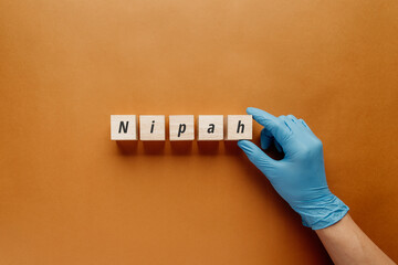 A medical professional's hand in a blue glove places wooden blocks spelling Nipah. This concept visualizes the Nipah virus outbreak, zoonotic infection, medical research, and disease prevention.