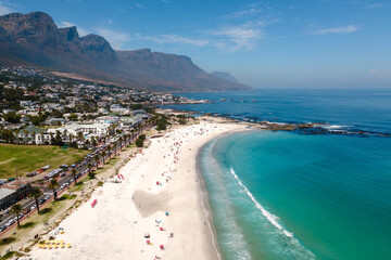Bright sun casts a golden glow on Camps Bay beach Cape Town, where visitors lounge on fine sand under colorful umbrellas. The calm azure water and majestic mountains create an idyllic vacation setting