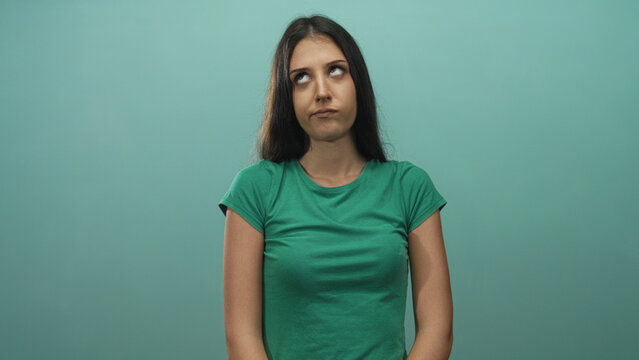 Young hispanic woman in green t shirt rolling eyes and pursing lips while standing and facing camera in studio with teal backdrop; annoyance.