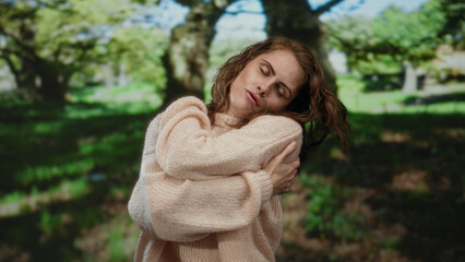 Young woman embraces herself contentedly in a serene park setting, surrounded by lush green trees, suggesting a moment of peaceful solitude and self-appreciation.