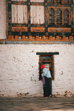 View of an elderly woman in traditional attire peers through an ornate window against a textured white wall, Jambay Lhakhang, Jakar, Bhutan.
