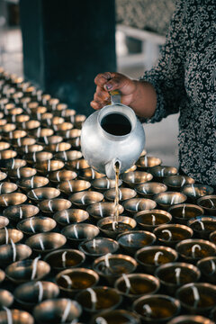 View of woman's hands pouring liquid from a silver kettle into rows of golden offering bowls, creating a serene scene, Jambay Lhakhang, Jakar, Bhutan.