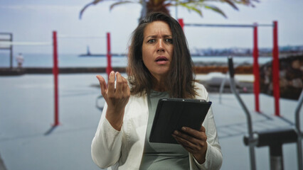 Woman holding tablet with raised hand gesture on street by outdoor fitness bars and sea  frustration confusion. © Krakenimages.com