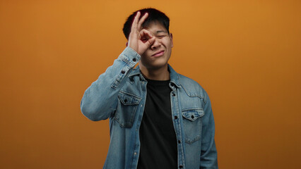 Fototapeta premium Young man wearing denim jacket making ok gesture over isolated orange background, showcasing confidence and communication.