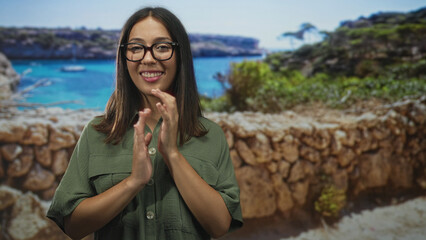 Fototapeta premium Young hispanic brunette woman clapping hands on a stone seaside promenade by turquoise water wearing green shirt and glasses; joy.