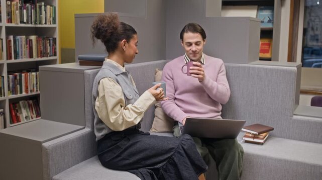 Medium long shot of young biracial girl bringing tea mugs to young Caucasian male student working on laptop in public library with stack of books nearby, helping him to work on thesis
