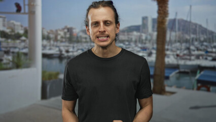 Man with long hair in black shirt clenches fists at marina pier, gritting teeth and leaning forward among moored boats and palm tree  anger resolve. © Krakenimages.com