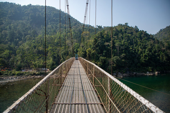 Shillong, India - 18 January 2019: View of the Dawki Bridge stretching across the Umngot River, connecting the land with a metal pathway, surrounded by lush green hills.