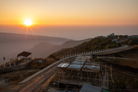 Shillong, India - 21 January 2019: View of a golden sun dipping below the horizon, casting warm hues over winding roads and distant structures.