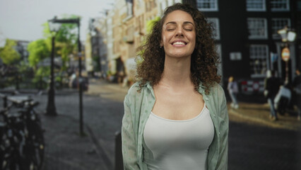 Fototapeta premium Woman smiling with eyes closed, wearing open shirt and white tank showing bare chest on a street with bicycles and lamp posts; calm contentment.