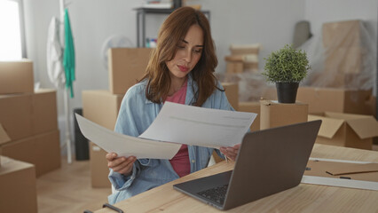 Woman working on laptop surrounded by moving boxes in new apartment, reviewing documents, green...