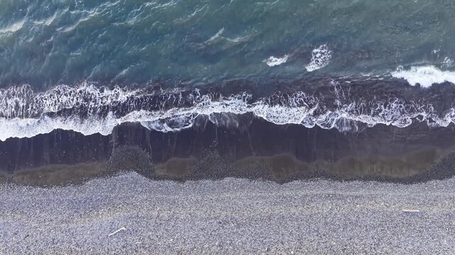 Top-down aerial drone footage starting with a close overhead view of ocean waves and foam patterns on a dark shoreline, followed by a rapid vertical ascent revealing a wider coastal surface.