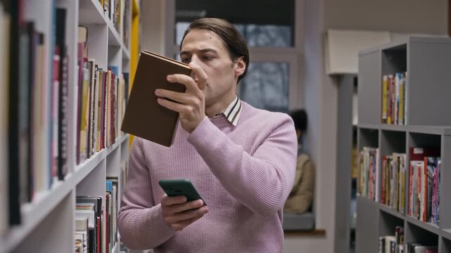 Medium shot of focused young Caucasian male student in smart casual outfit standing in library aisle holding smartphone and searching for books working on his thesis or dissertation