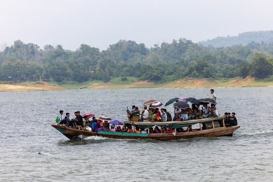 Rangamati, Bangladesh - 18 June 2023: View of a bustling wooden boat, its vibrant passengers a splash of color against the serene backdrop of lush green hills and shimmering water.
