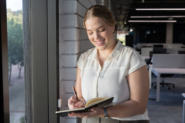 Woman writing notes in notebook by window next to brick column in open-plan office