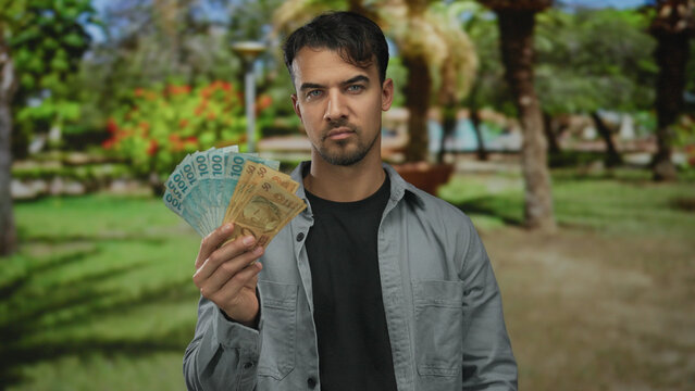 Young hispanic man holding brazilian banknotes thoughtfully standing in a lush outdoor park with tropical trees and vibrant greenery in the background.