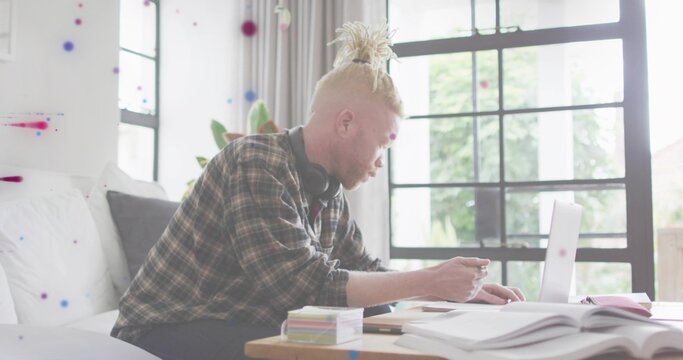 Sitting man wearing plaid flannel shirt and headset, working in living room with laptop, notebooks