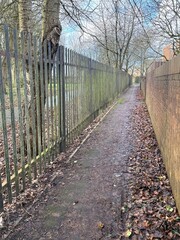 Narrow pedestrian path between metal fence and brick wall with fallen leaves, quiet urban walkway scene in late autumn or early winter with bare trees.