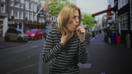 Fototapeta premium Woman coughing into fist with hand on throat on a city street, striped top visible; illness discomfort.
