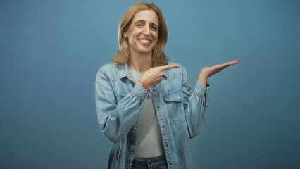 Fototapeta premium Woman points finger to palm while smiling in blue studio backdrop wearing denim jacket and gray shirt; friendly presentation.