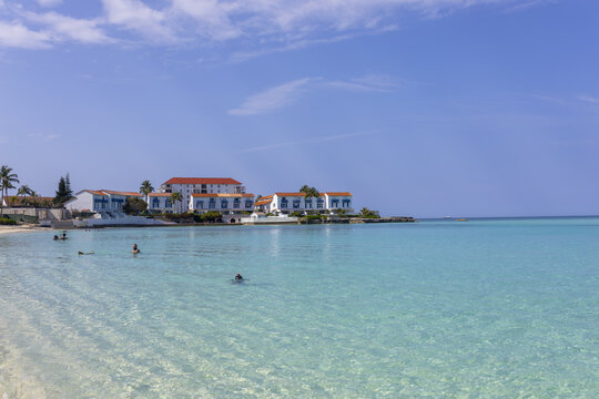 View of turquoise water gently laps against the sandy beach, reflecting the clear blue sky near white buildings, Nassau, New Providence, The Bahamas.