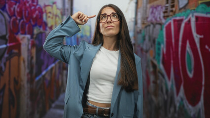 Naklejka premium Woman with glasses points finger to temple in street alley with colorful graffiti walls; thoughtful curiosity.